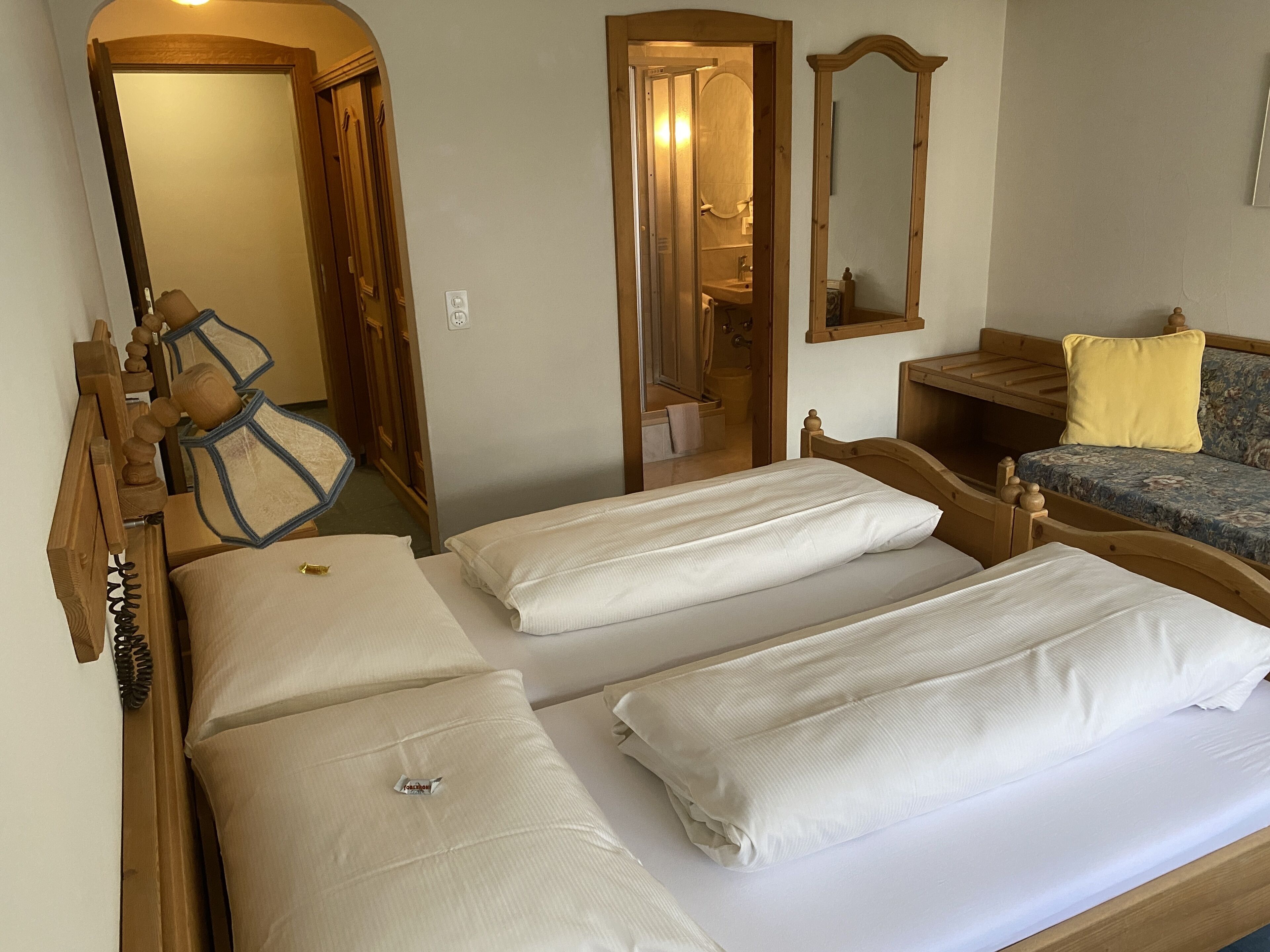A room with two beds, a wooden headboard, a mirror, a small sofa with a cushion, and a wooden bench.