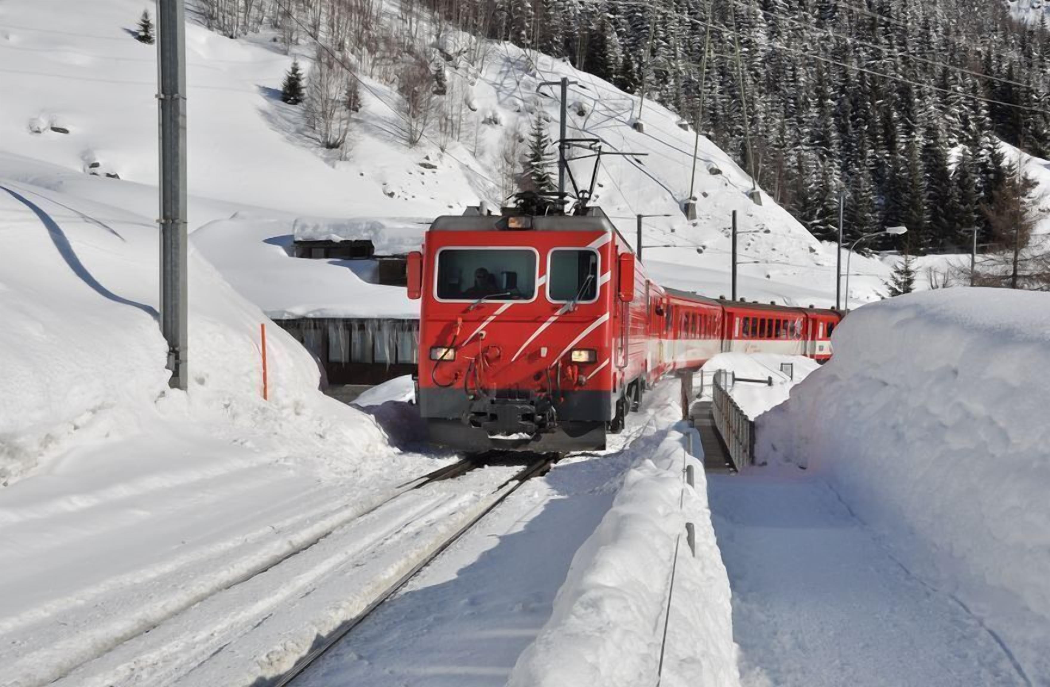 Un tren rojo sobre una vía cubierta de nieve, con montañas al fondo.