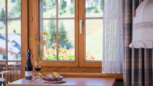 A wooden table with a wine bottle, a glass, and a plate with food and flowers by a window with a view of greenery and buildings. - B&B Garni Casa La Val (Tujetsch)