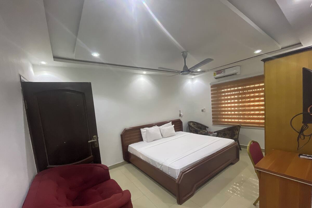 A bedroom with a bed, a red chair, a desk, a ceiling fan, and a window with blinds.