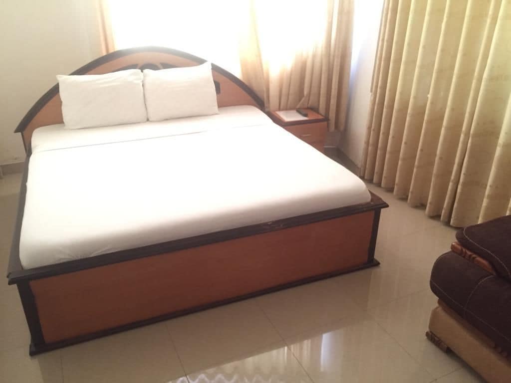 Una cama grande con estructura de madera y ropa de cama blanca, una mesita de noche con lámpara y una ventana con cortinas.
