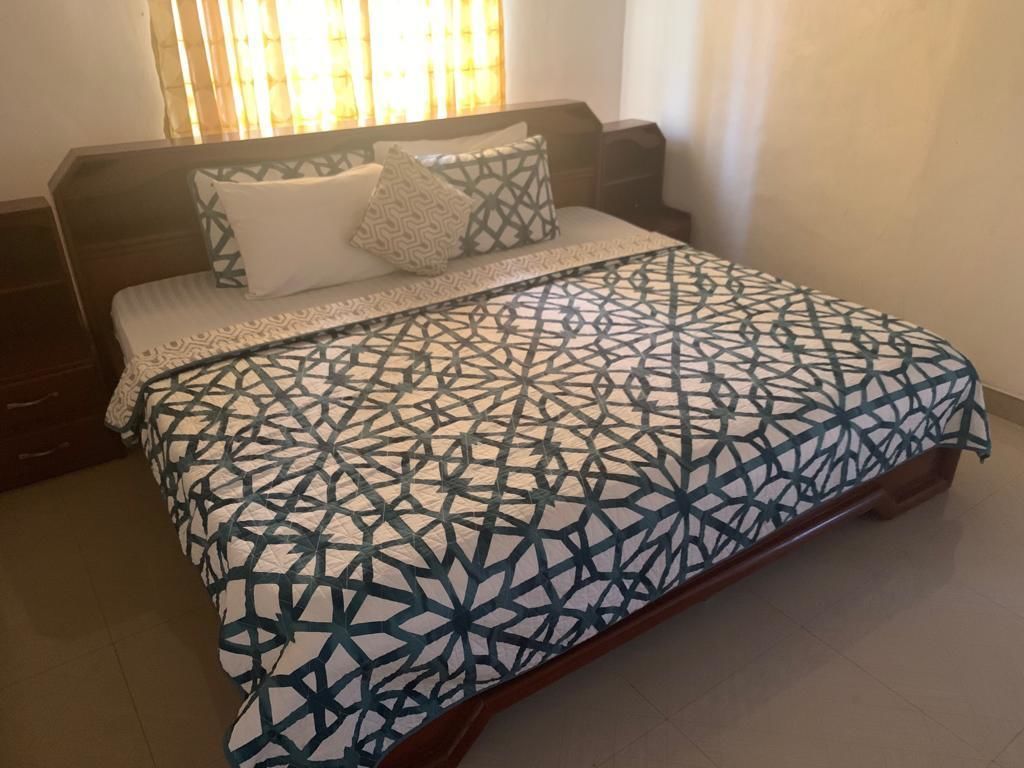 A bed with a geometric patterned bedspread, a wooden headboard, and a bedside table with a lamp.