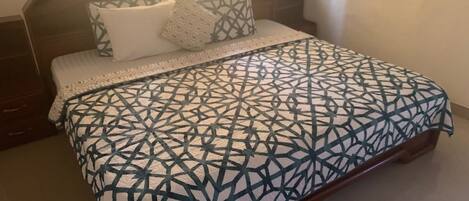 A bed with a geometric patterned bedspread, a wooden headboard, and a bedside table with a lamp.
