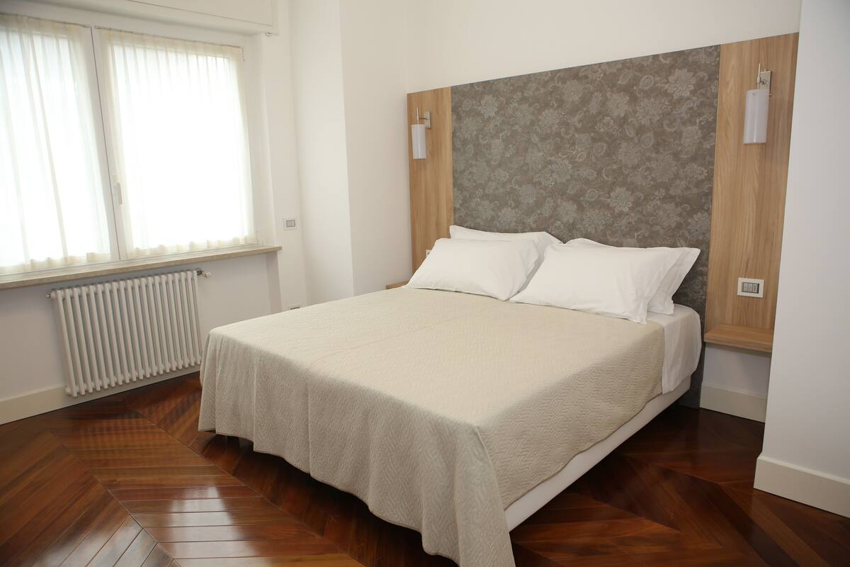 Ein ordentlich bezogenes Bett mit einer beige farbenen Tagesdecke in einem Raum mit Holzboden und einer Heizung.