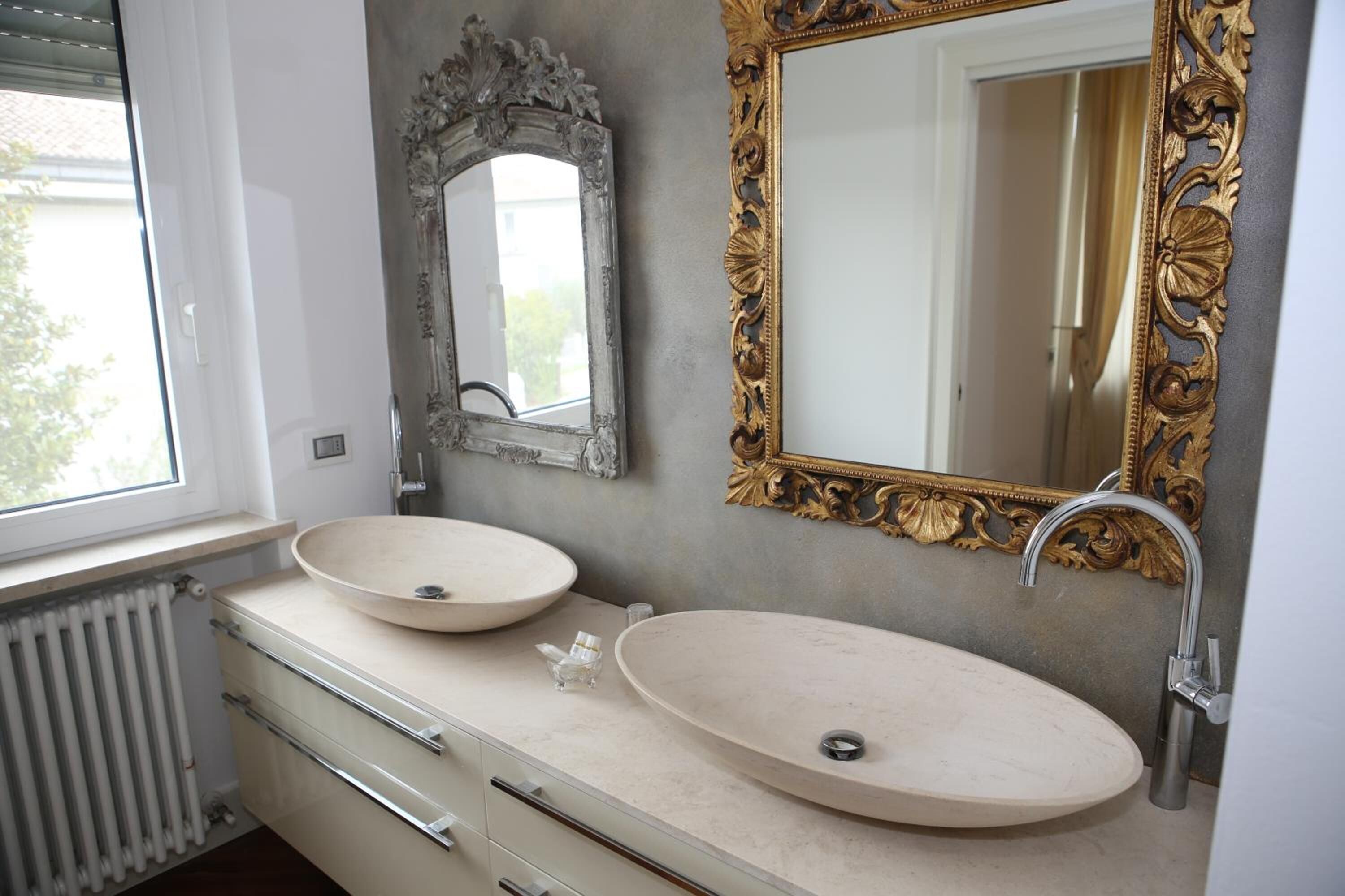 A bathroom with two oval sinks, a window, a radiator, and two ornate mirrors.