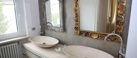 A bathroom with two oval sinks, a window, a radiator, and two ornate mirrors.