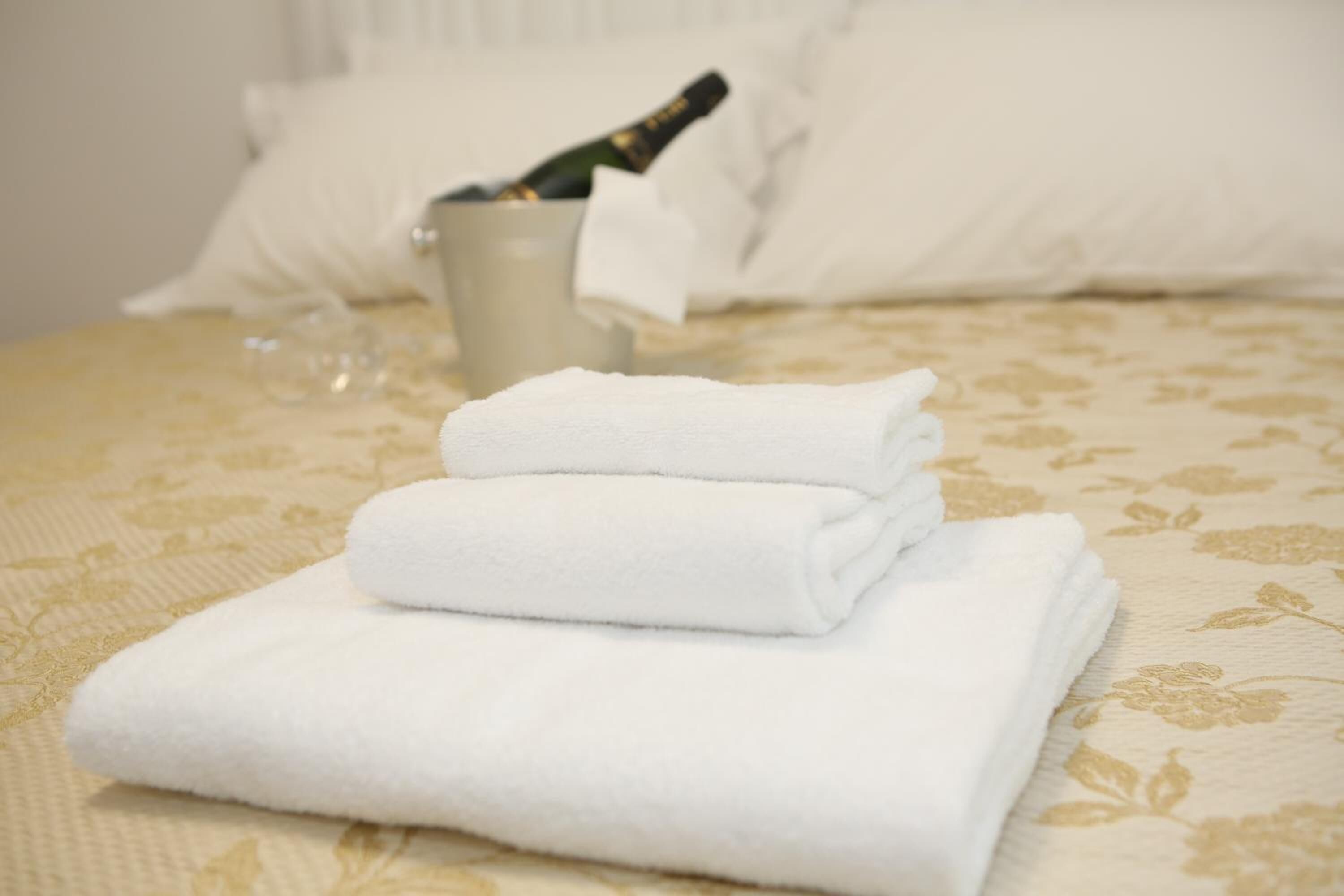 A neatly made bed with folded white towels and a bottle of champagne in a glass.