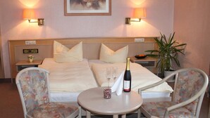 Hotelkamer met een bed, twee bedlampen, een schilderij, een nachtkastje, een potplant en een tafel met een fles en glazen.