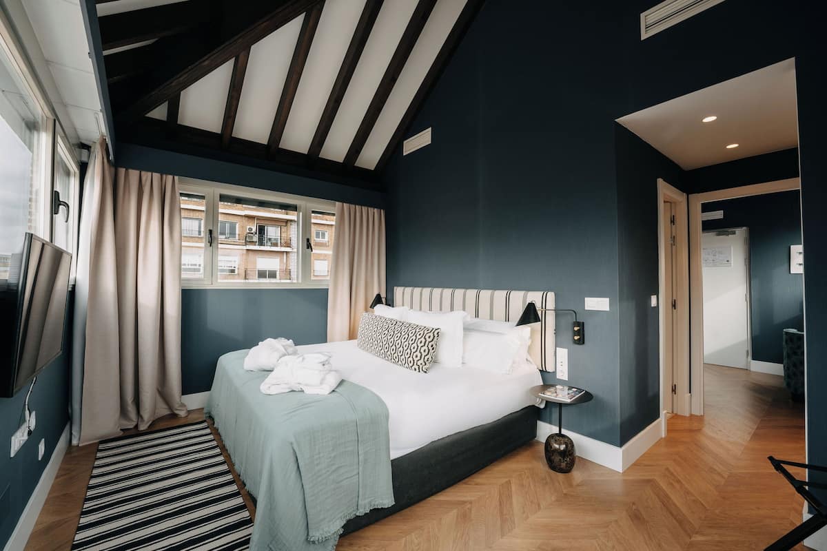 A bedroom with a large bed, a bedside table, a TV mounted on the wall, and a window with curtains.