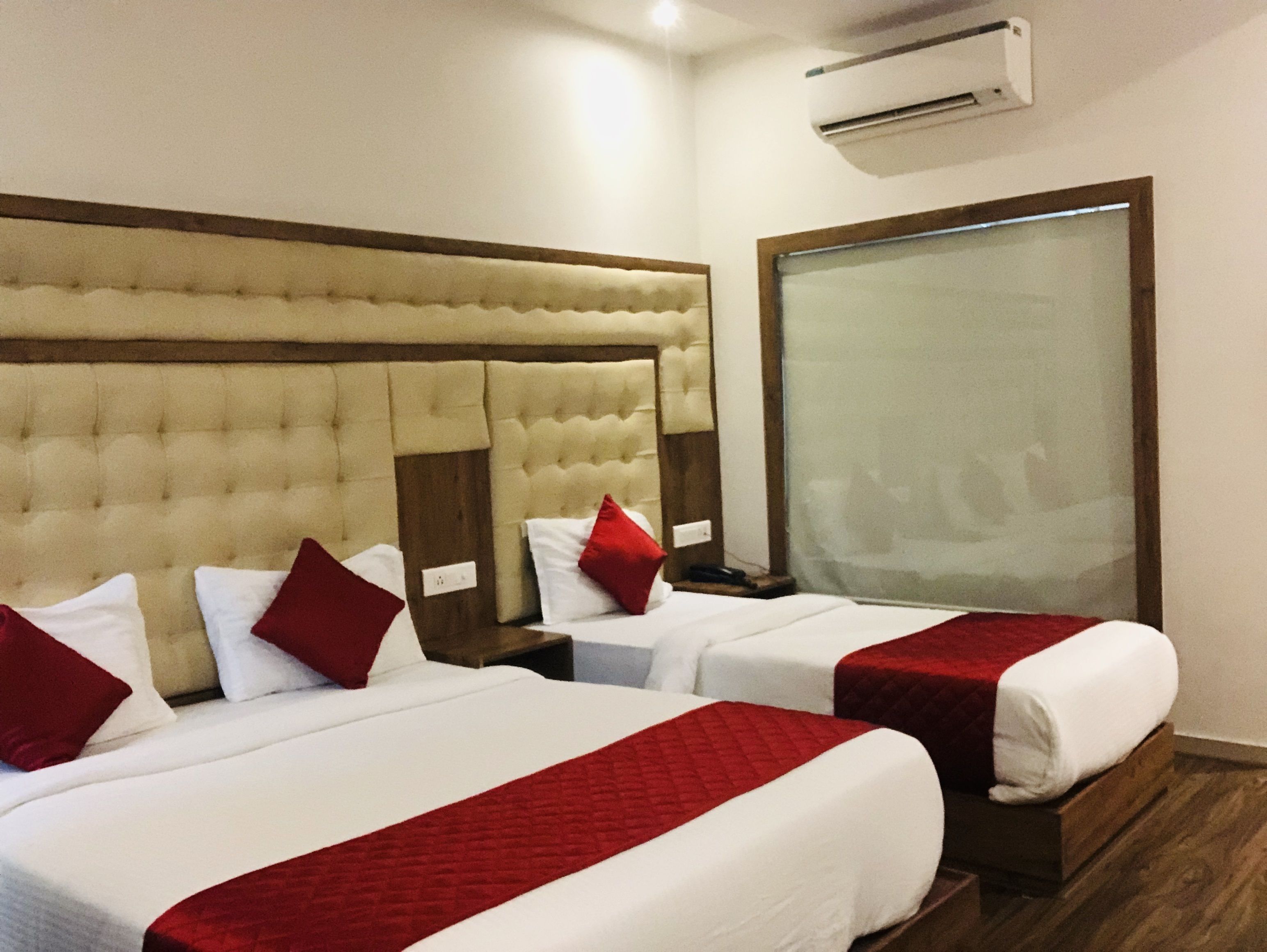 A hotel room with two beds, each with red and white bedding, a large window, and an air conditioning unit on the wall.