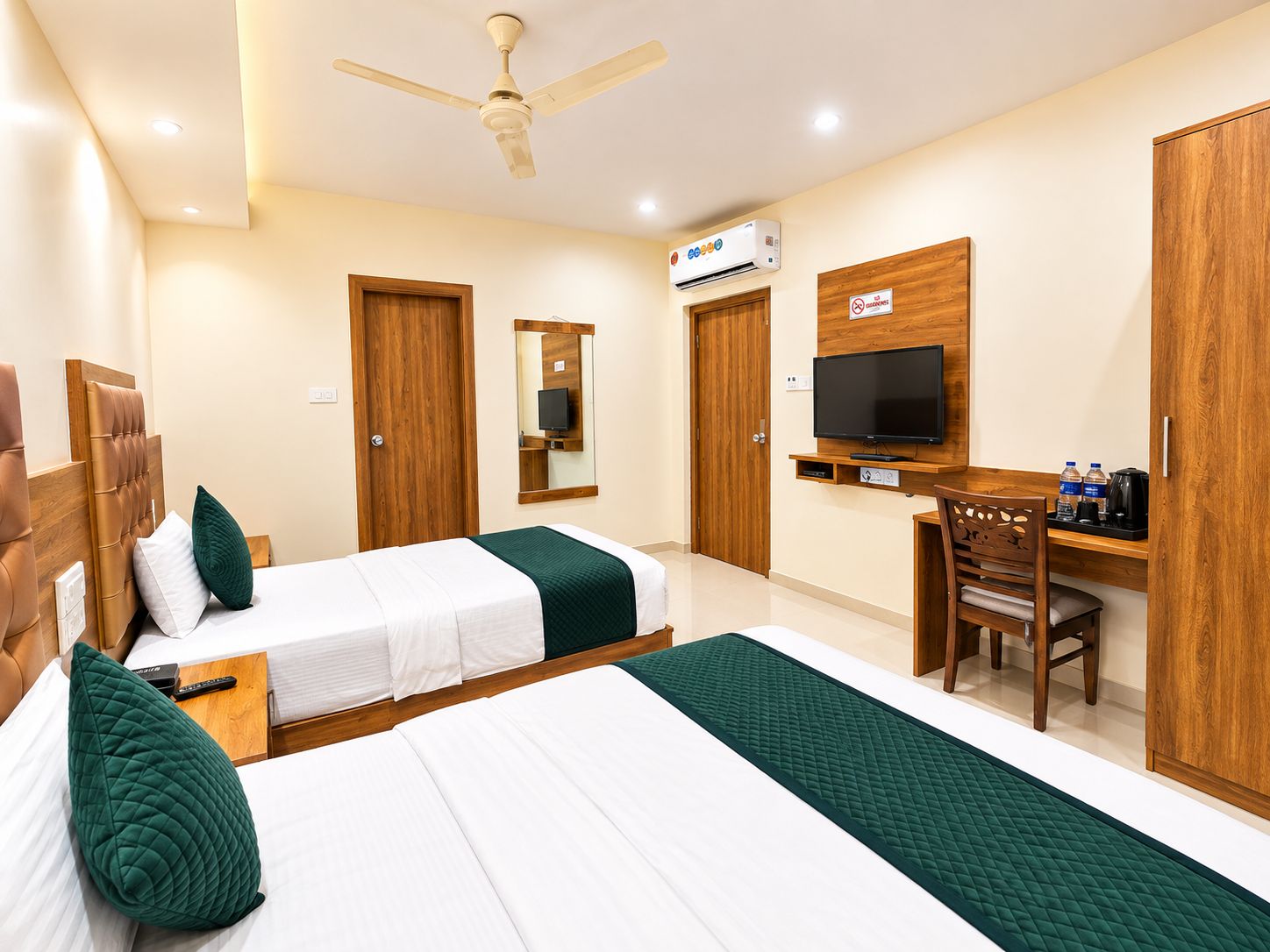 A hotel room with two beds, a desk, a television, and a ceiling fan.
