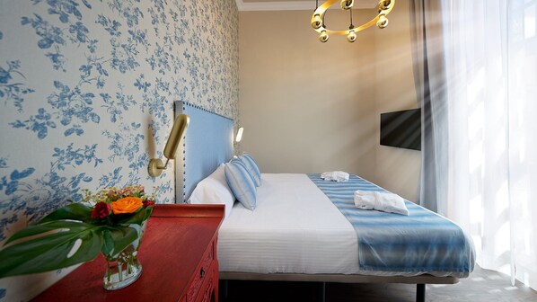 Quarto de hotel com cama, mesa de cabeceira com um vaso de flores e televisão fixada na parede.