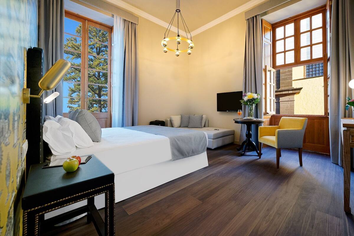 Habitación de hotel con cama, mesita de noche, lámpara y una pequeña mesa con un florero.