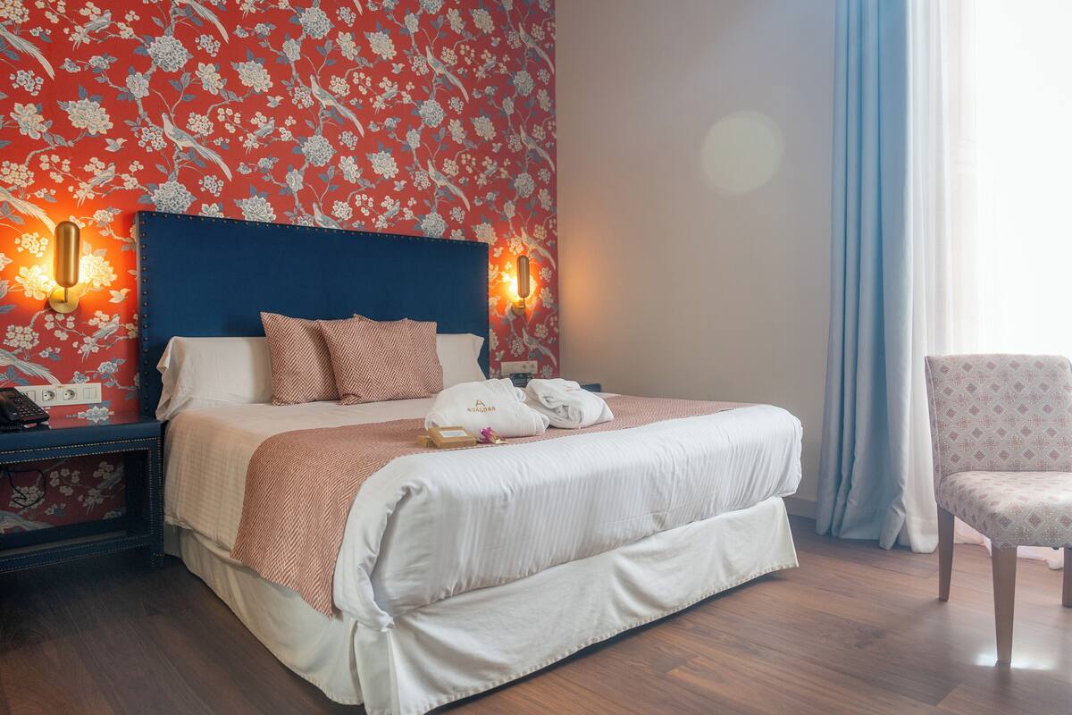 Una cama bien hecha con un cabecero azul, una mesita de noche con un teléfono y una lámpara, y una silla con un cojín estampado.