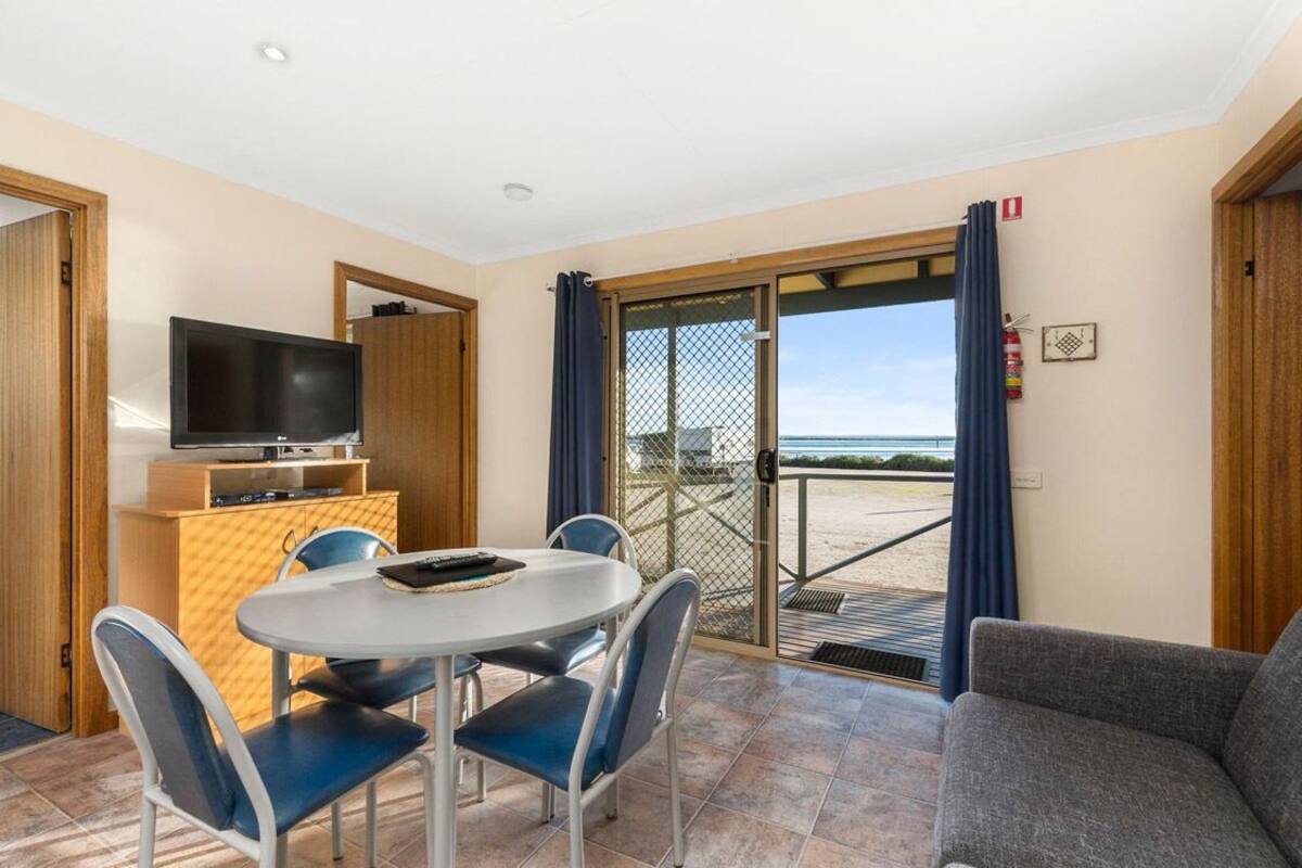 Een kamer met een televisie, een eettafel met stoelen, een bank en uitzicht op het strand door een glazen deur.