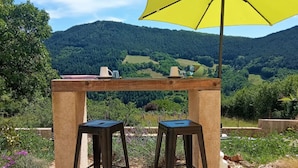 A wooden table with two stools and a yellow umbrella, set against a backdrop of a lush valley and forested hills. - Chambres d'hôtes & Spa La Villa des Raspes - Adults Only (Viala-du-Tarn)