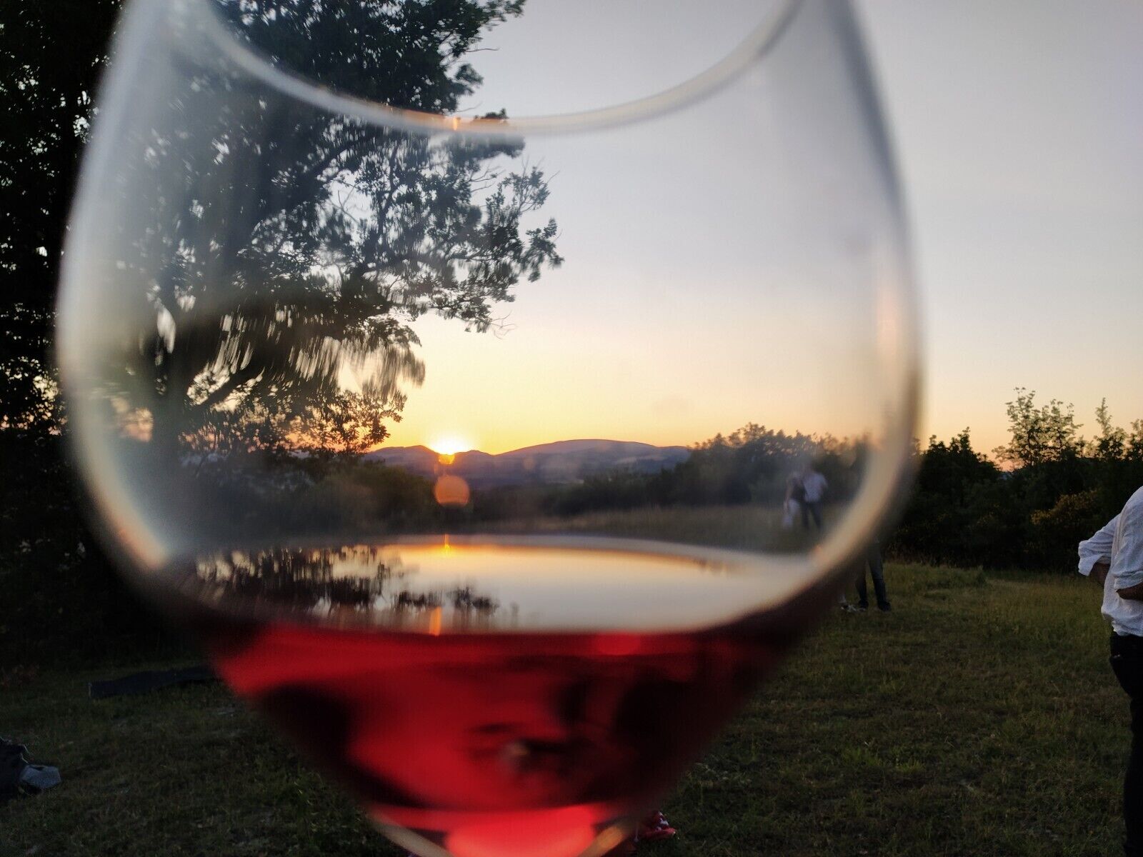 A glass of red wine with a reflection of a sunset and a person in the background.