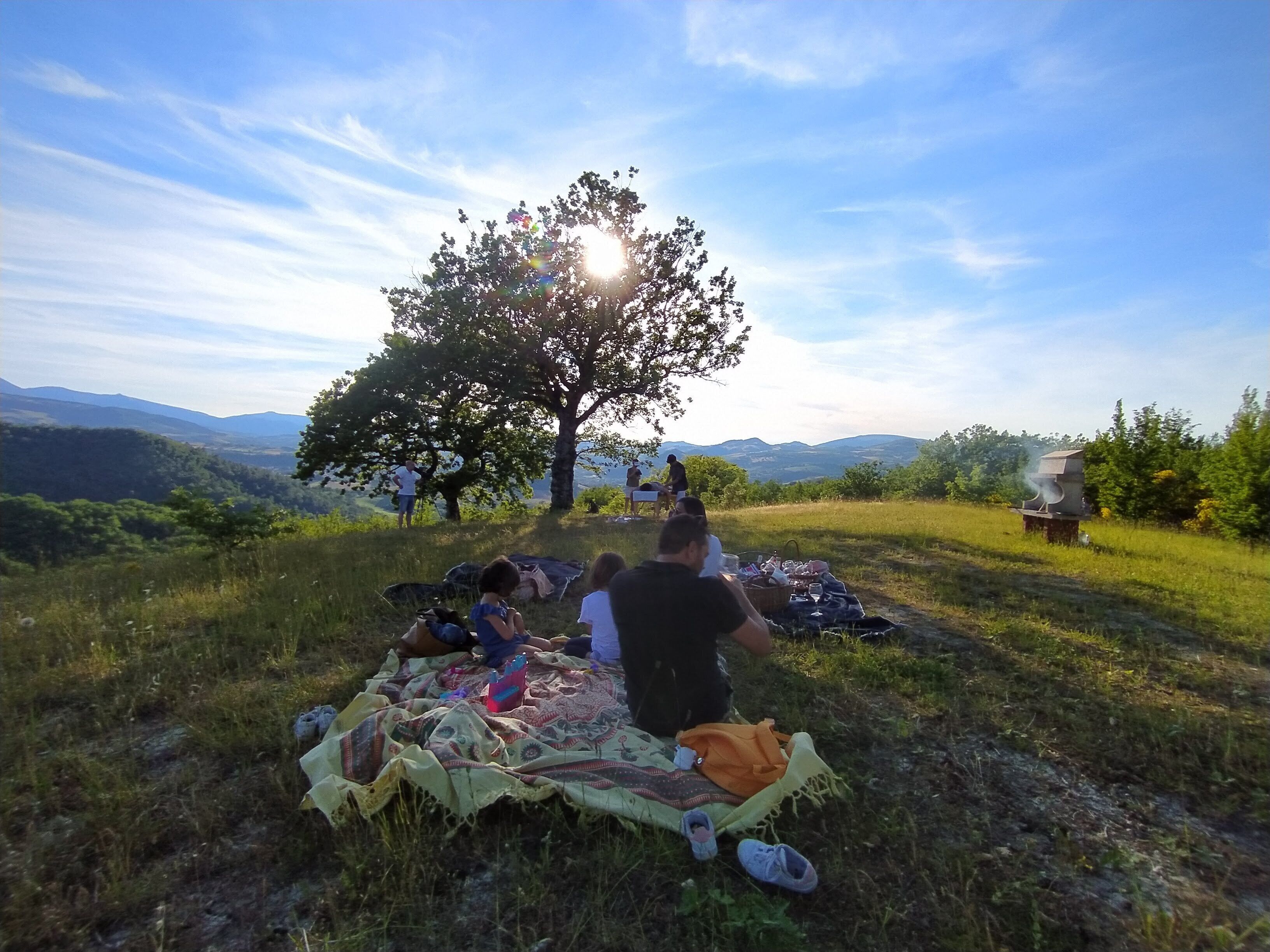 Eine Gruppe von Personen sitzt draußen auf einer Decke, im Hintergrund sind ein Baum und ein Grill zu sehen.