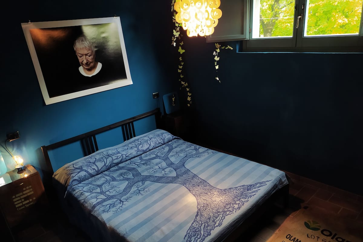 A bedroom with a bed, a bedside table, a wall-mounted picture, a window with a view of greenery, and a hanging light fixture.