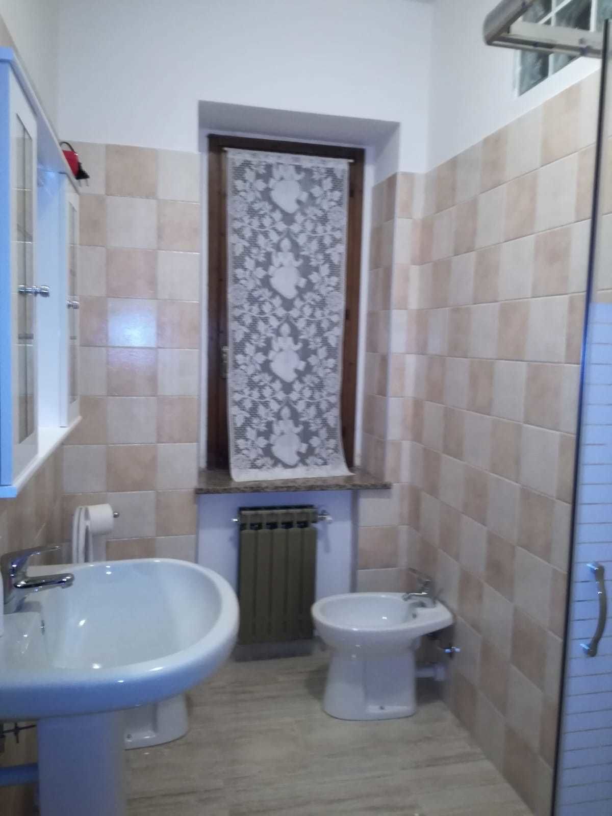 Un baño pequeño con lavamanos, inodoro y bidé. Hay un perchero sobre el inodoro y un radiador debajo.