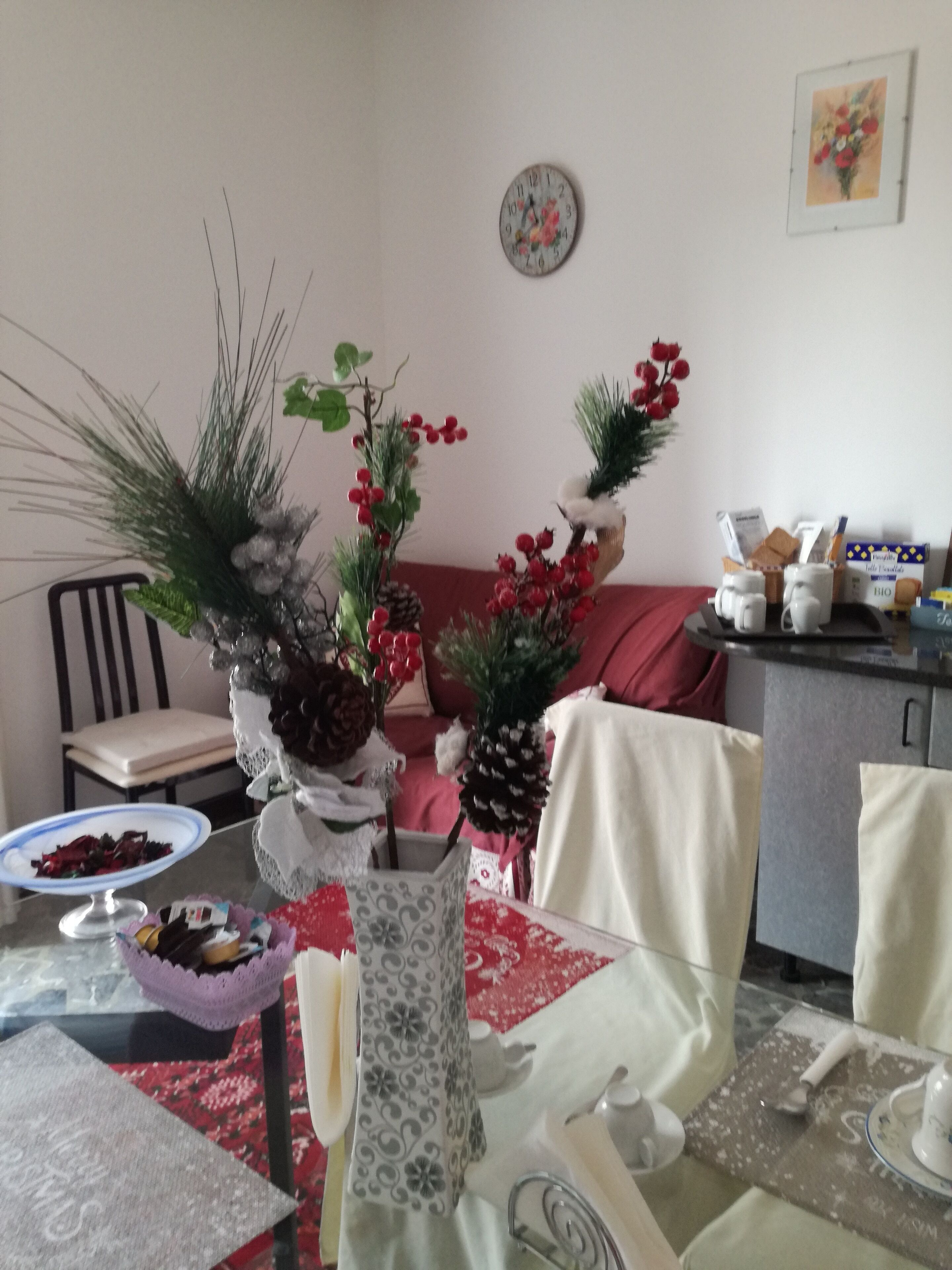 Un comedor con una mesa dispuesta para dos personas, con un centro de mesa decorativo que incluye ramas de pino y bayas rojas. Se ve un reloj en la pared, y hay un pequeño cuadro sobre la mesa.