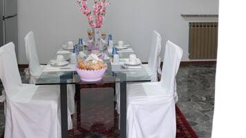 Un coin repas avec une table en verre, des chaises blanches, un tapis à motifs rouges, un vase avec des fleurs roses et un luminaire au plafond.