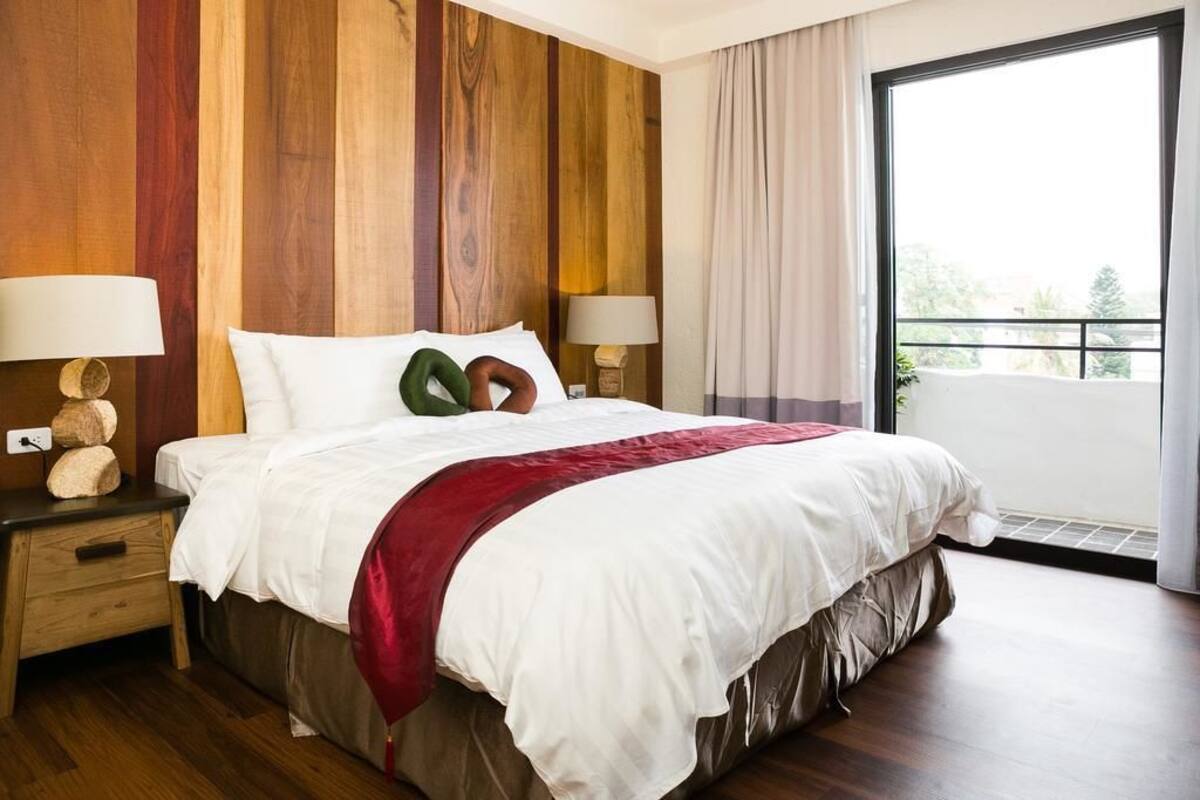 A hotel room with a large bed, wooden panel walls, two bedside tables with lamps, and a view of greenery through a large window.