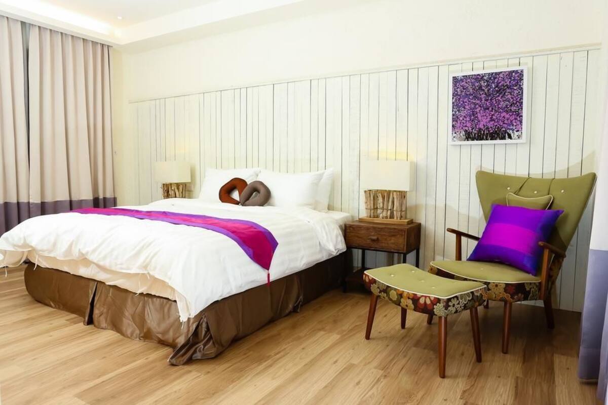 A hotel room with a large bed, two bedside tables, a green armchair, a small table, and a purple pillow.