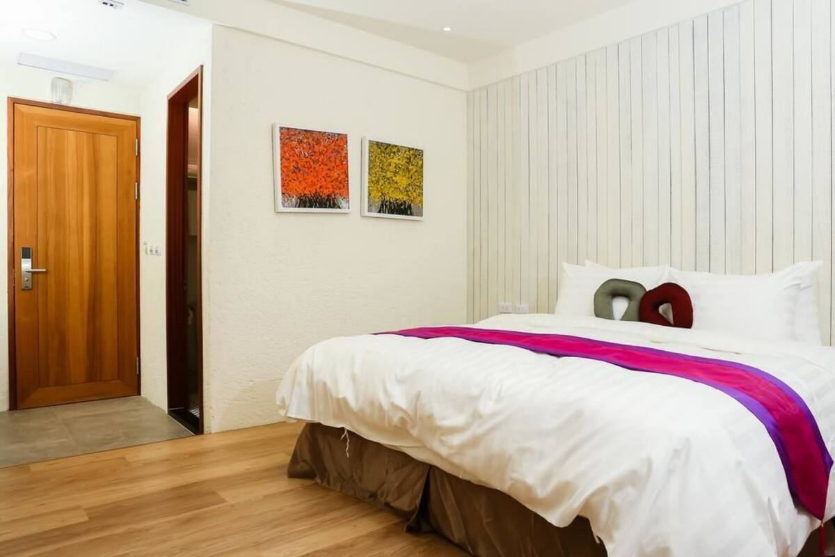 A bedroom with a wooden door, a bed with white bedding and a pink accent, and two wall-mounted paintings.