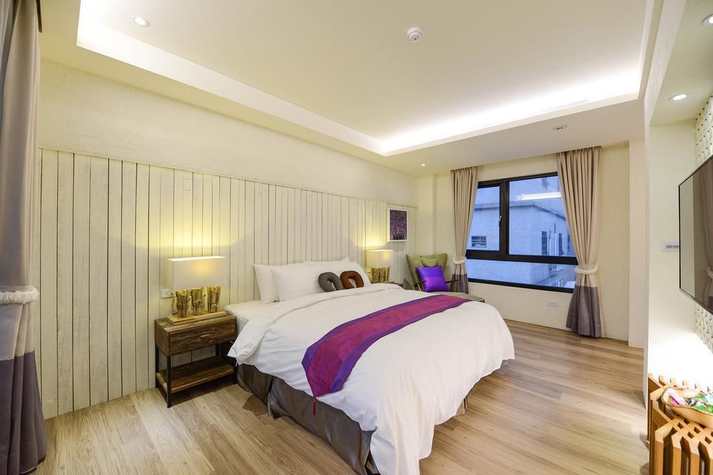A modern hotel room with a large bed, wooden flooring, a nightstand with a lamp, a window with curtains, and a TV on the wall.