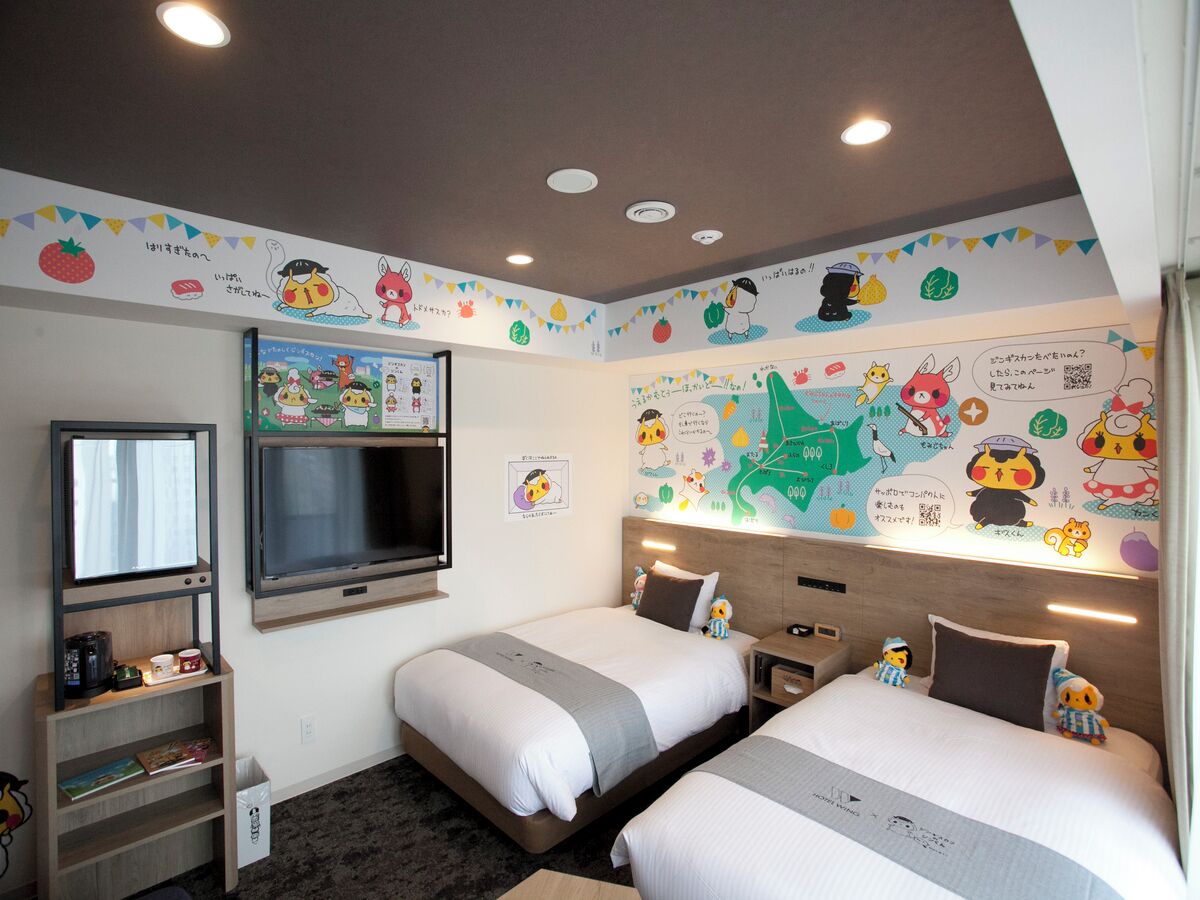 Une chambre d’hôtel avec deux lits, une télévision, une étagère remplie de livres et un mur décoré de personnages de dessins animés.