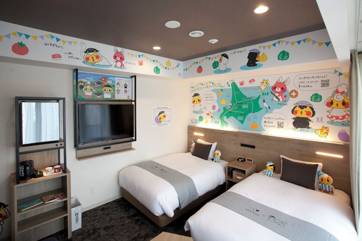 A hotel room with two beds, a TV, a shelf with books, and a wall decorated with cartoon characters.