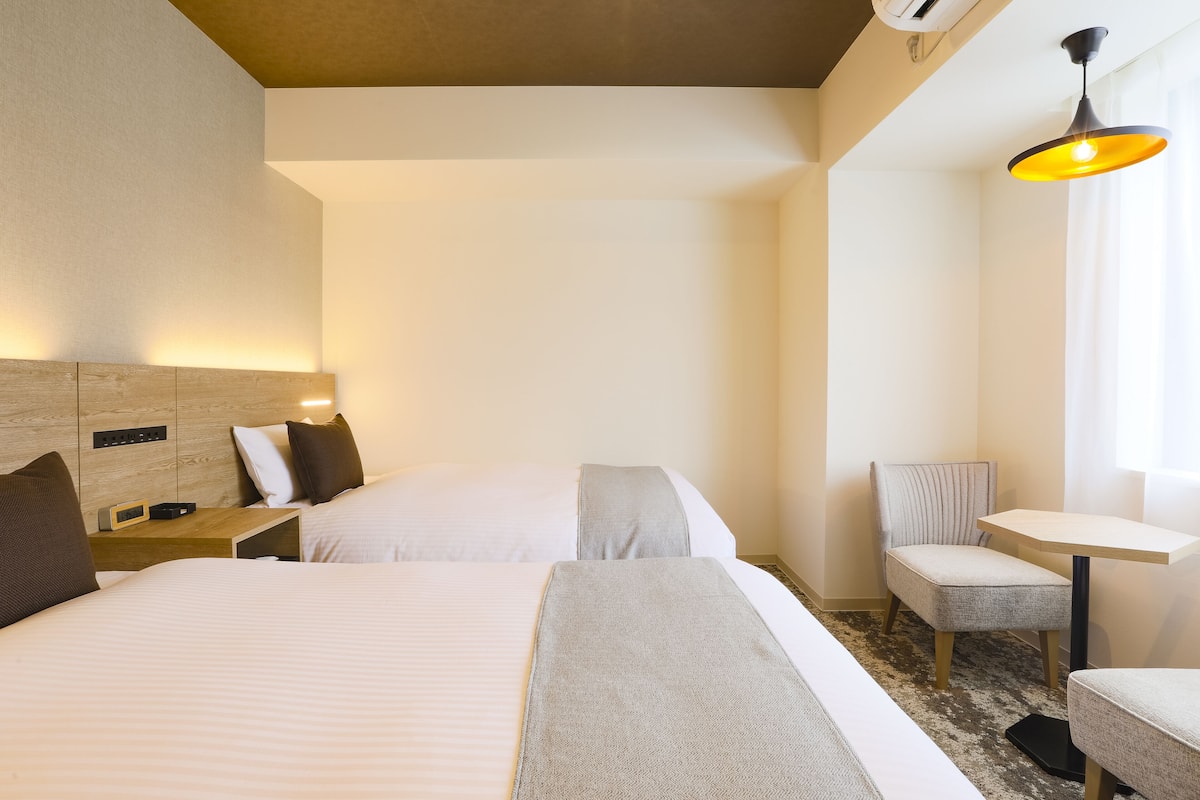 A hotel room with two beds, a small table, a chair, and a bedside table with a lamp.