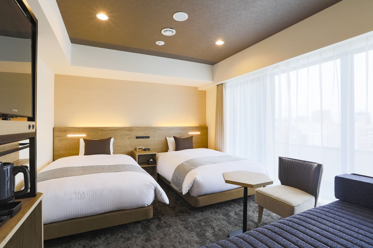A hotel room with two beds, a small table, a chair, a coffee maker, and a large window with curtains.