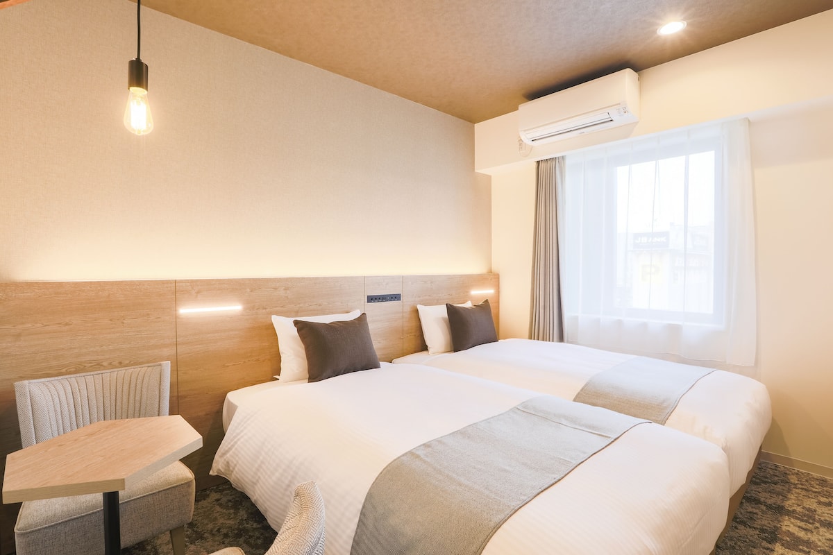 A hotel room with a large bed, a small table, a chair, a wall-mounted air conditioner, and a window with curtains.