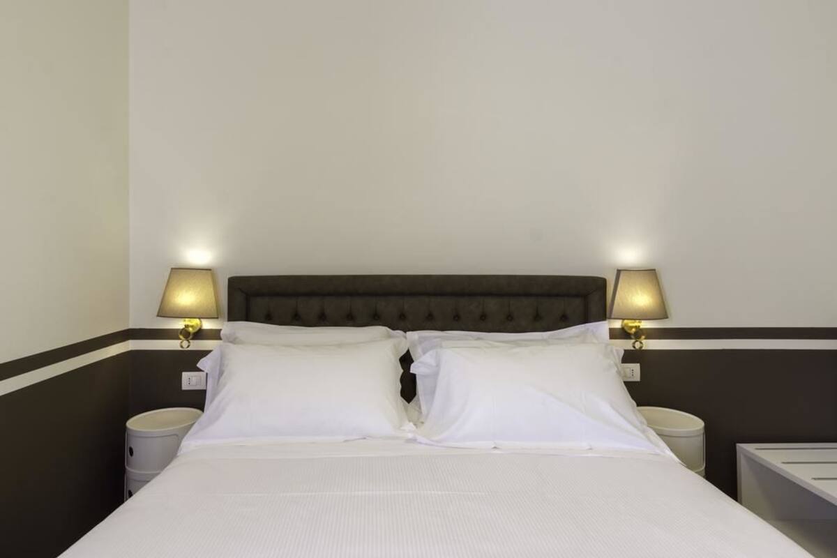 Un lit bien fait, avec du linge de lit blanc, deux lampes de chevet et une tête de lit sombre.