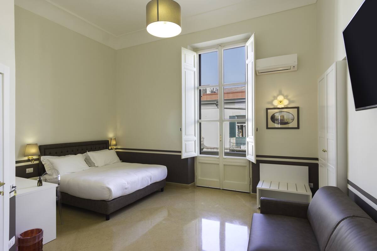 Une chambre d’hôtel avec un lit, des tables de chevet, un canapé, une fenêtre avec une porte ouverte et un téléviseur fixé au mur.