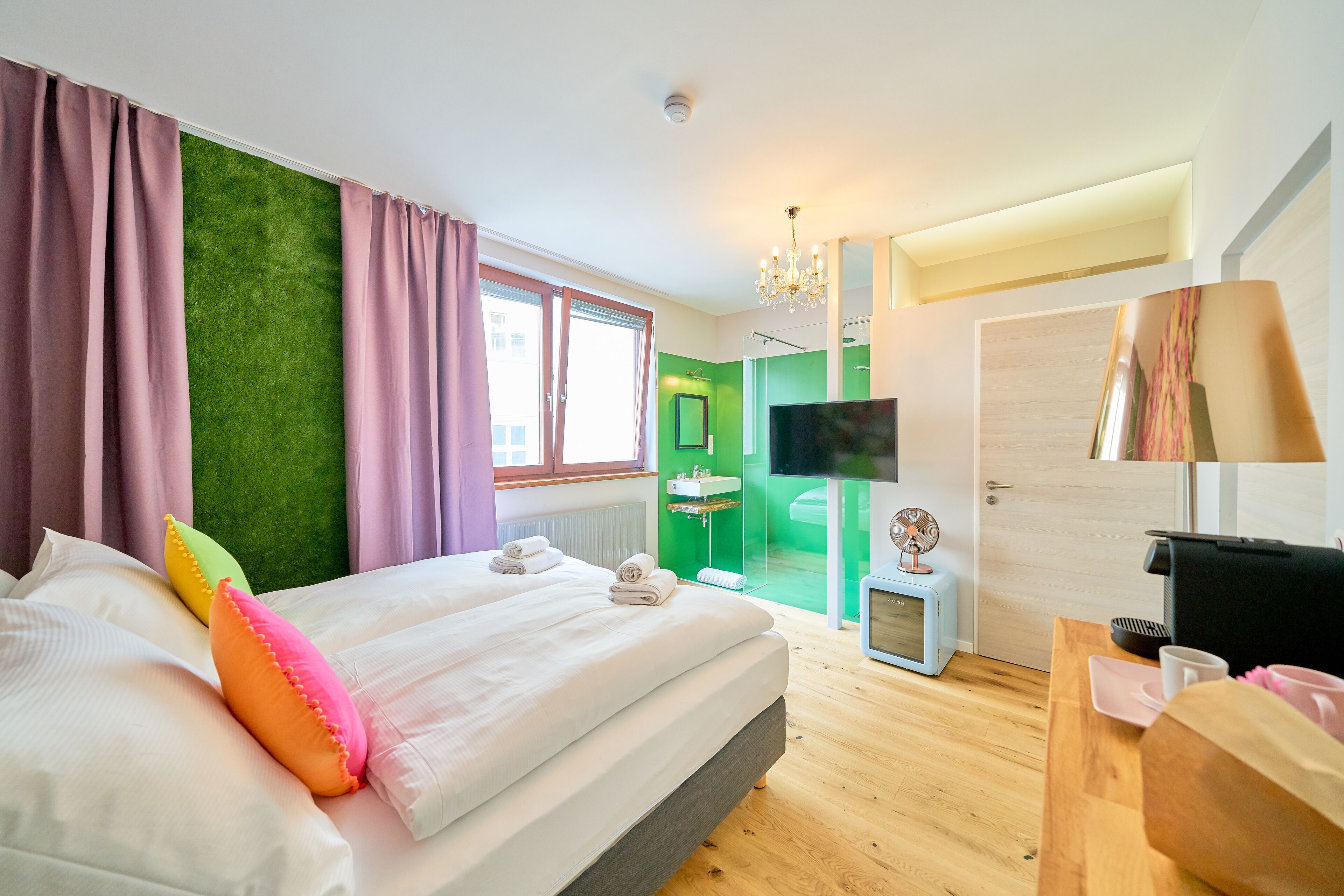 A modern hotel room with a large bed, a television, a desk with a coffee maker, and a green accent wall.