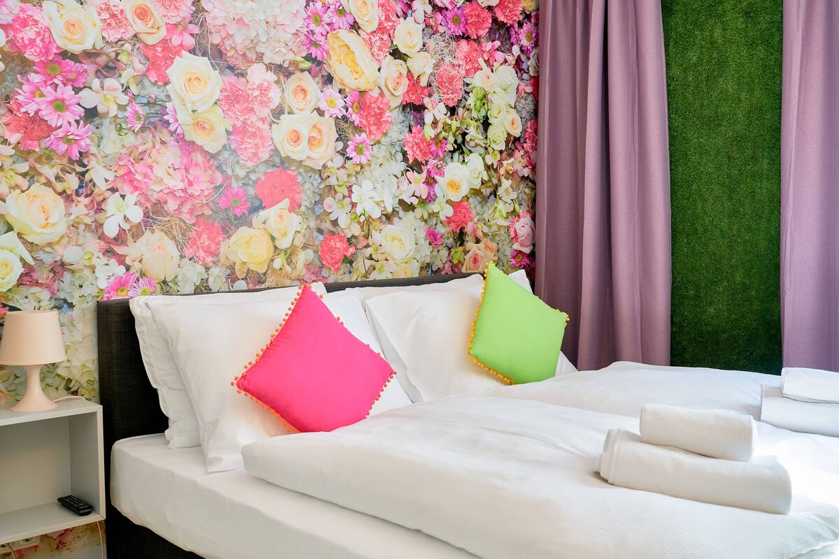 A bed with white linens, two pillows, a remote control on a nightstand, and a floral wallpaper backdrop.