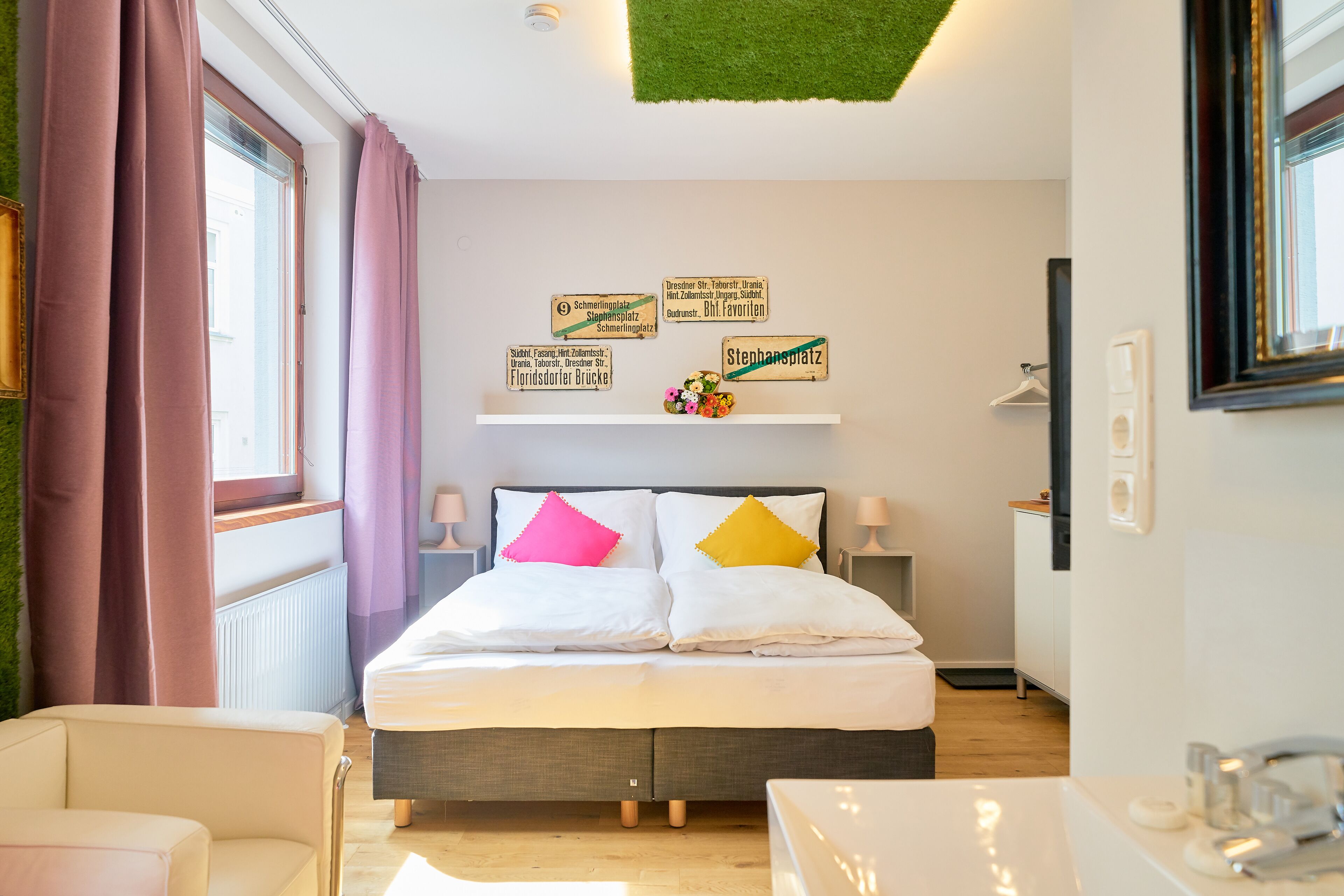 Habitación de hotel con cama, una silla, una mesa y una ventana con cortinas. Hay carteles en la pared que indican la ubicación.