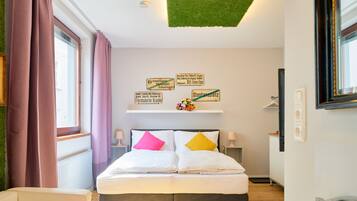 Habitación de hotel con cama, una silla, una mesa y una ventana con cortinas. Hay carteles en la pared que indican la ubicación.