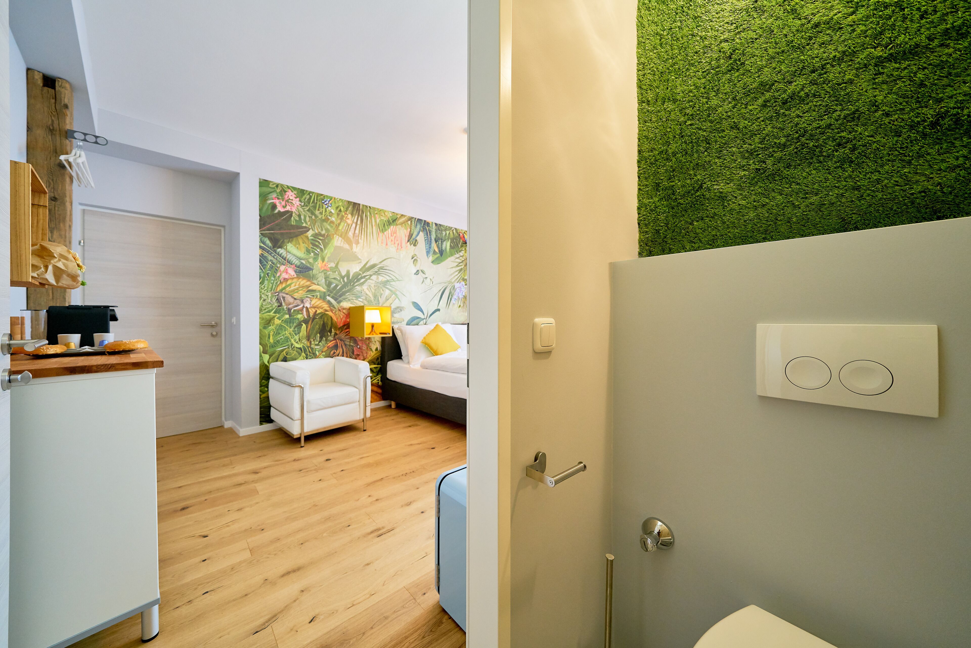 Ein modernes Hotelzimmer mit Holzboden, einer weißen Couch, einem Bett und einem Wandbild mit grünen Motiven. Eine Badezimmertür ist sichtbar.