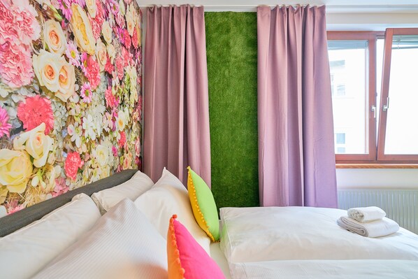 Habitación de hotel con un papel tapiz floral, un panel de césped artificial verde y una cama con ropa de cama blanca y almohadas coloridas.