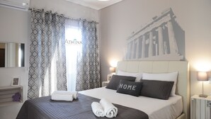 Une chambre d’hôtel comprenant un lit, une tête de lit, une table de chevet, une lampe, un mur orné d’une illustration représentant un bâtiment, et une fenêtre avec des rideaux à motifs.