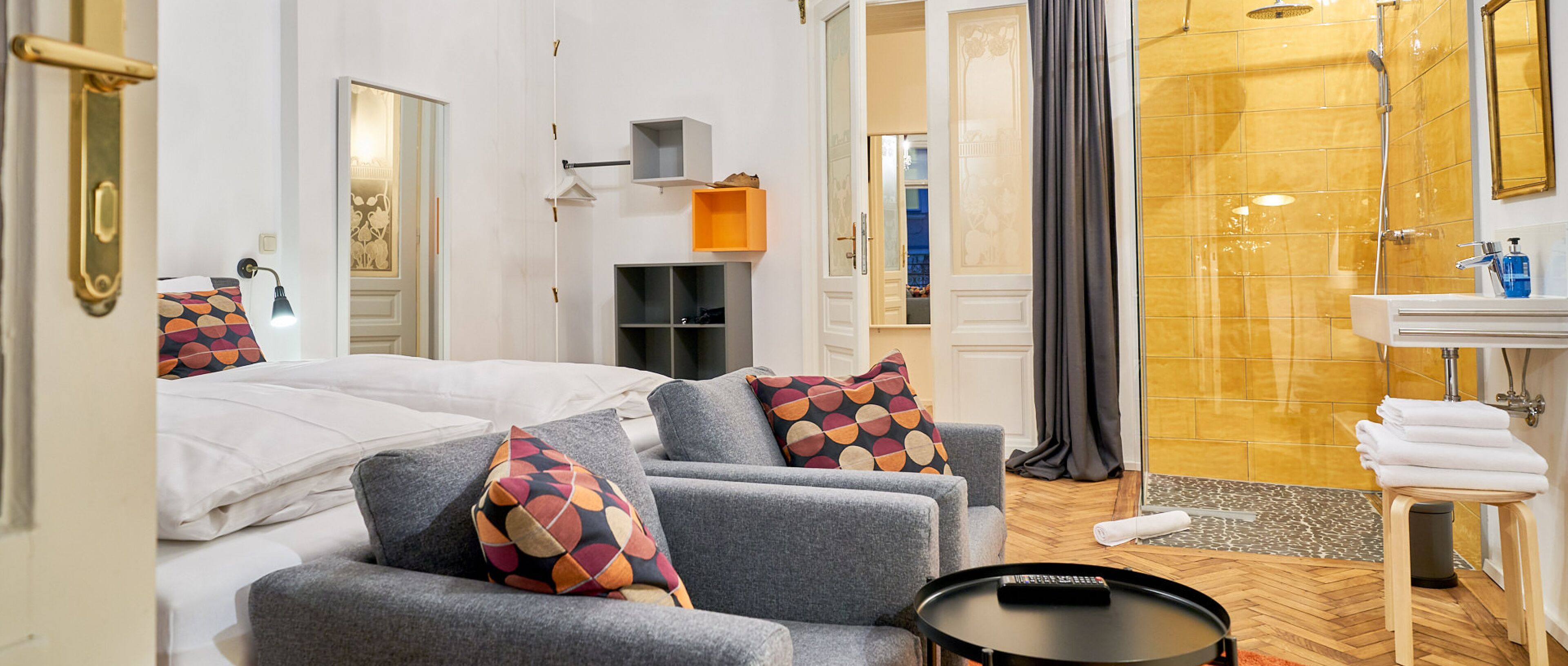 Ein Hotelzimmer mit Bett, grauem Sofa, bunten Kissen, einem kleinen Tisch, einer Duschkabine und einem Badezimmer mit Waschbecken und Handtuchhalter.