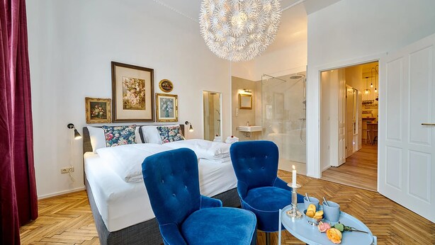 A modern hotel room with a large bed, two blue armchairs, a small round table with a candle, and a chandelier.