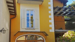 Een gebouw met twee verdiepingen, een geel geveloppervlak, een balkon met smeedijzeren hekwerk en een begane grond ingang met een houten deur en glaspanelen.