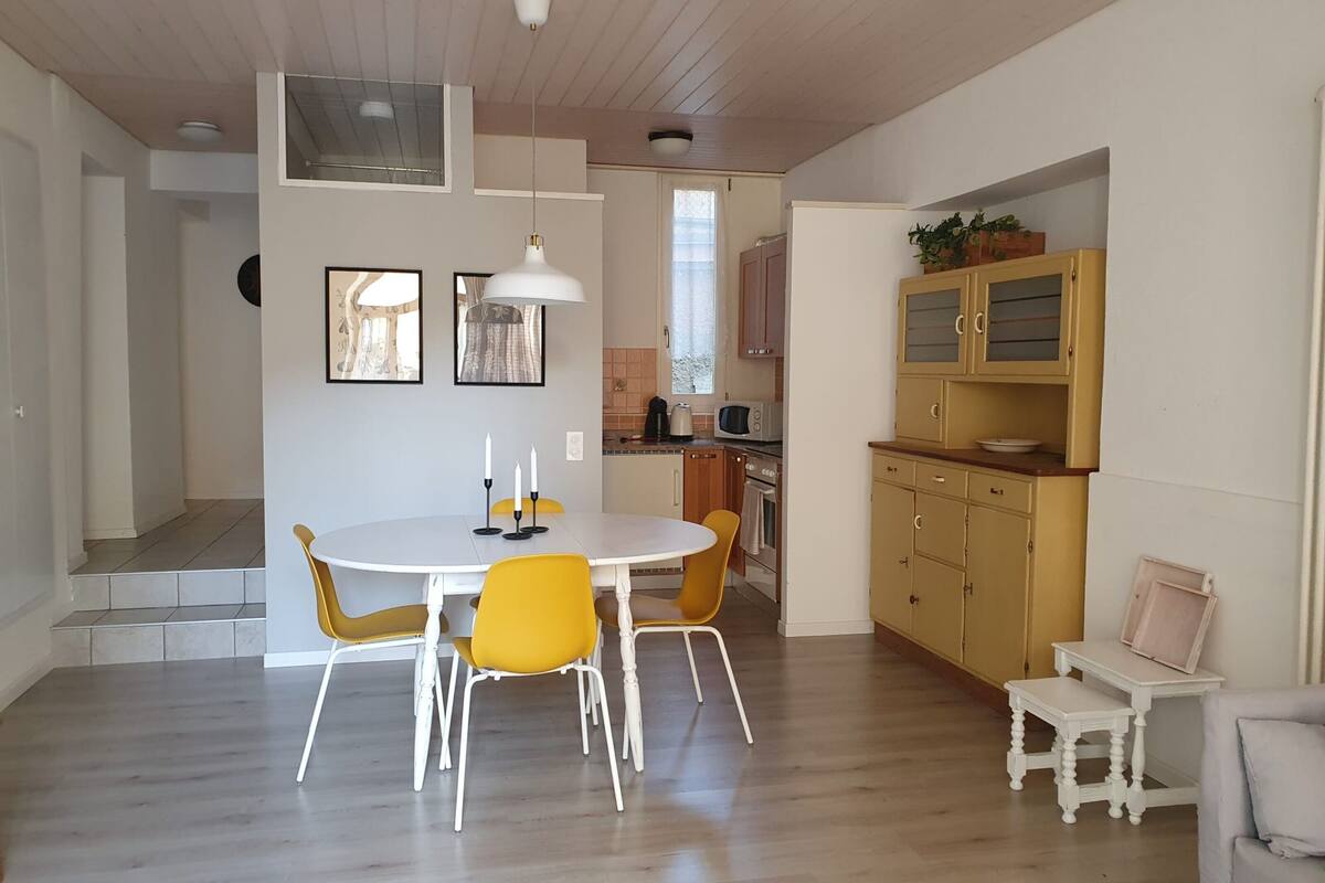 Eine moderne Küche mit Holzausstattung, ein Essbereich mit weißem Tisch und gelben Stühlen sowie ein kleiner weißer Tisch mit Bilderrahmen.