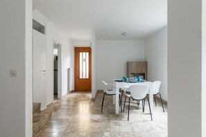 A narrow hallway with a dining area at the end, featuring a table and chairs, a wooden door, and a view into another room.