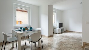 Een lichte, ruime kamer met een witte tafel en stoelen, een televisie en een raam.